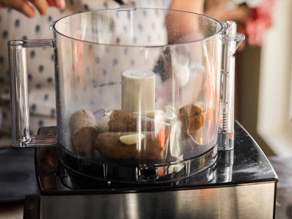 A person about to blend ingredients in a food processor. The bowl contains peeled garlic cloves and pieces of brown vegan sausage. The processor rests on a countertop, and a person is visible in the background wearing a polka dot shirt.